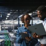 Data center system administrators using laptop to review AI LLM visualizations. African american IT specialists use device to analyze neural networks outputs, ensuring stable gear operations
