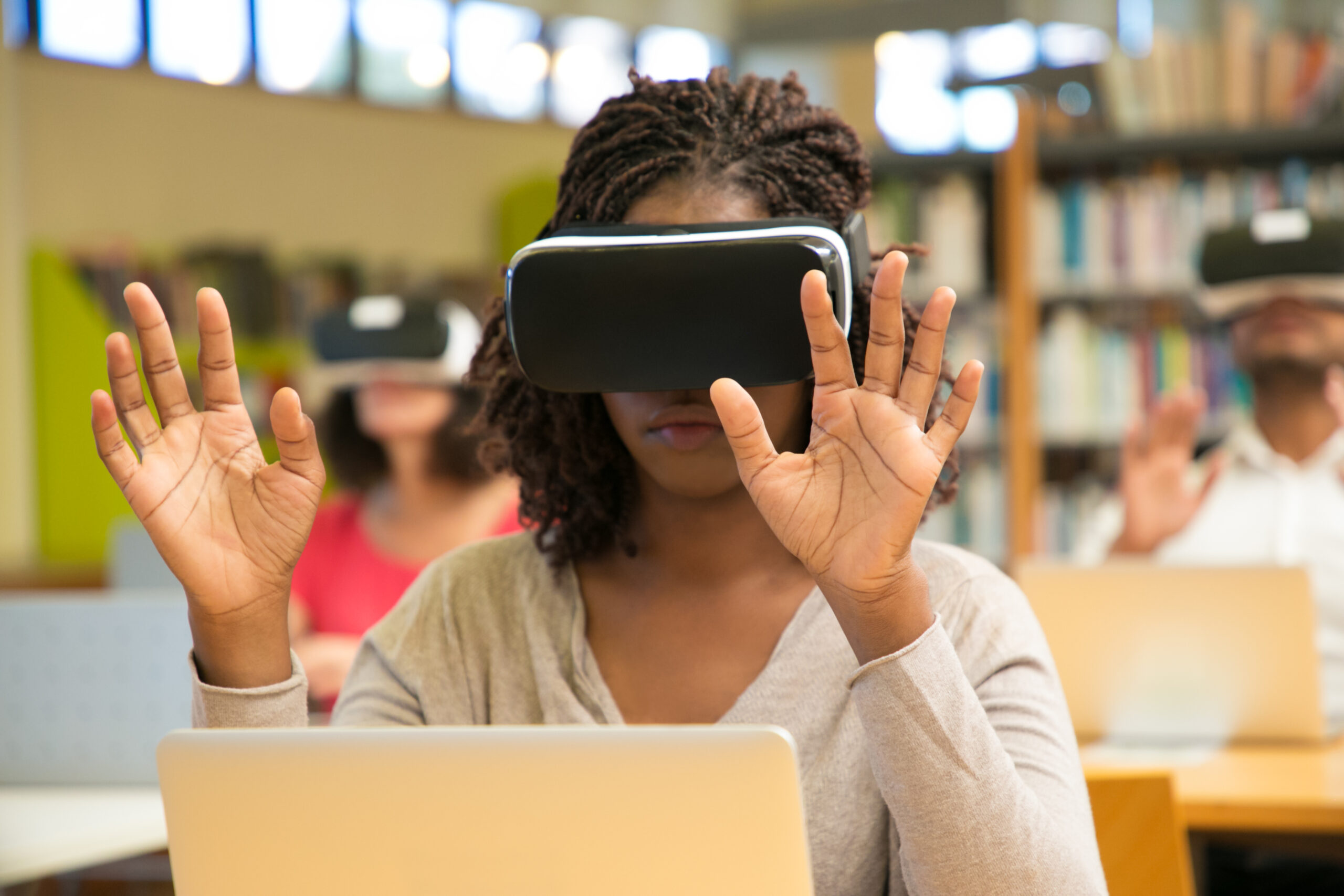 Multiracial group of students using VR gadgets during class. Young black woman in virtual reality headset sitting at desk with laptop and touching air. Devices for education concept.